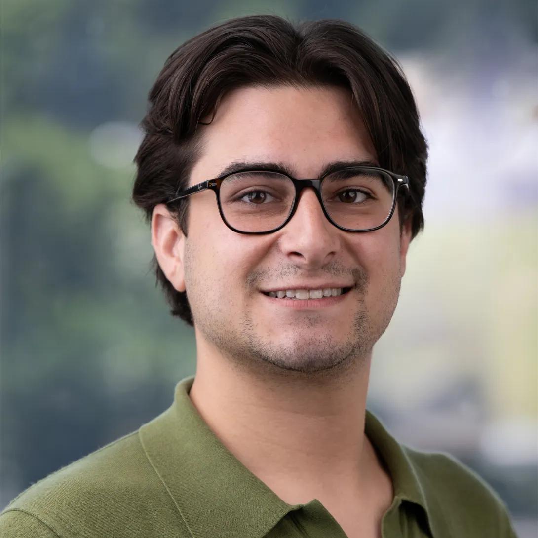 Head-and-shoulders portrait of a smiling man with dark, center-parted hair, light facial hair, and black rectangular glasses, wearing an olive-green collared shirt against a softly blurred background.