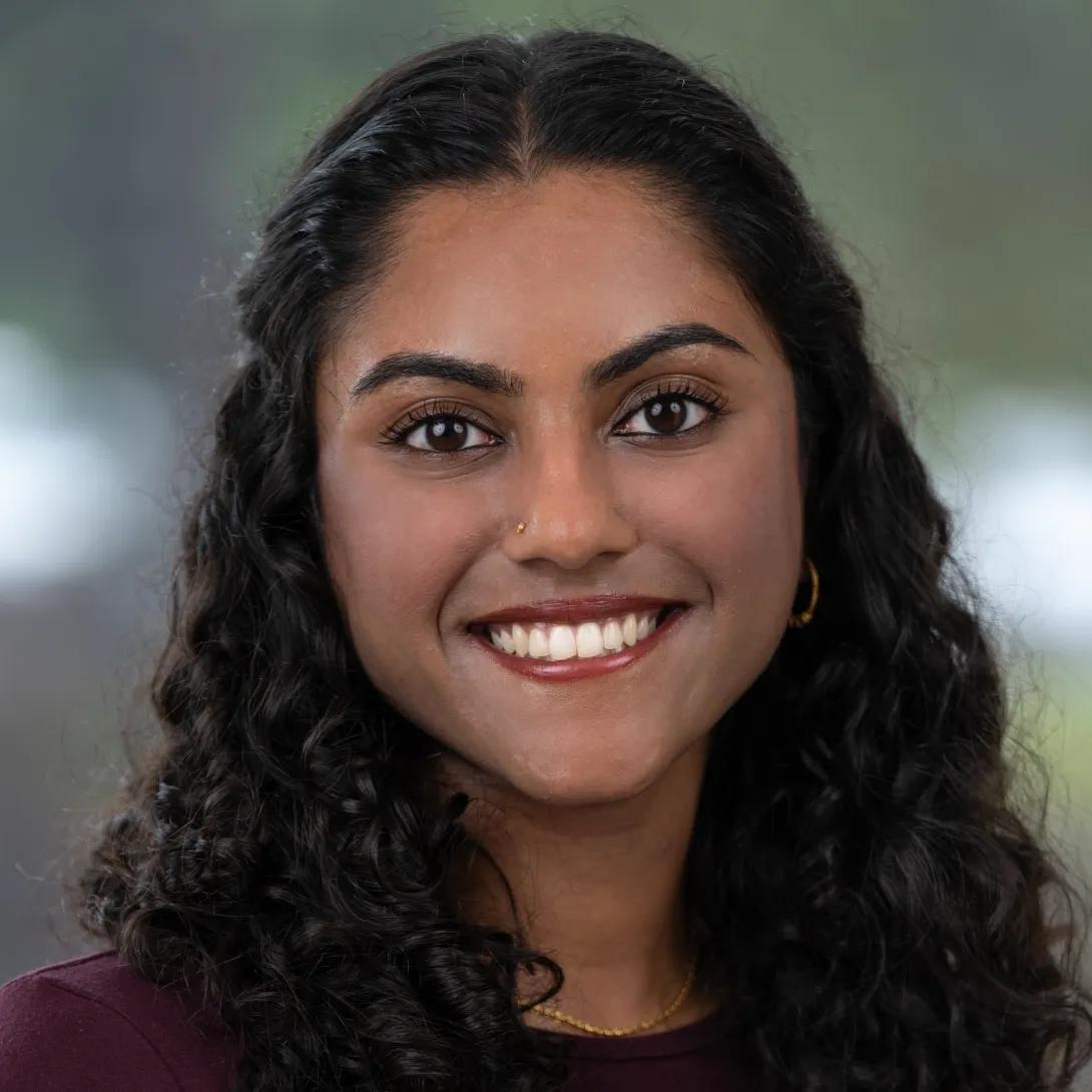 Professional headshot of a smiling woman with long, dark curly hair, a small gold nose stud, and gold jewelry, wearing a burgundy top against a softly blurred background.
