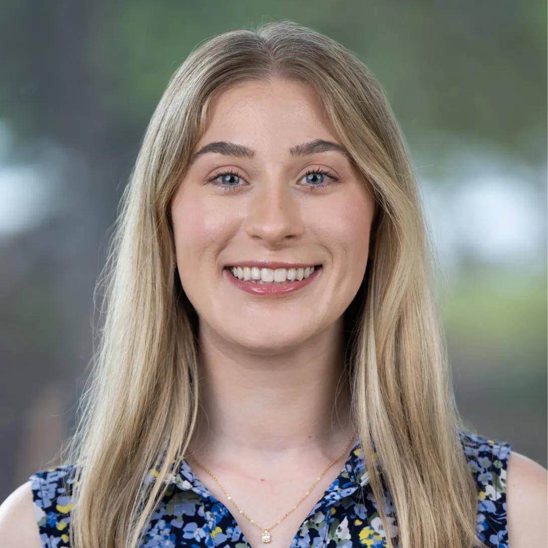 Professional headshot of a smiling woman with long, straight blonde hair, wearing a blue floral top and delicate gold necklace against a softly blurred background.