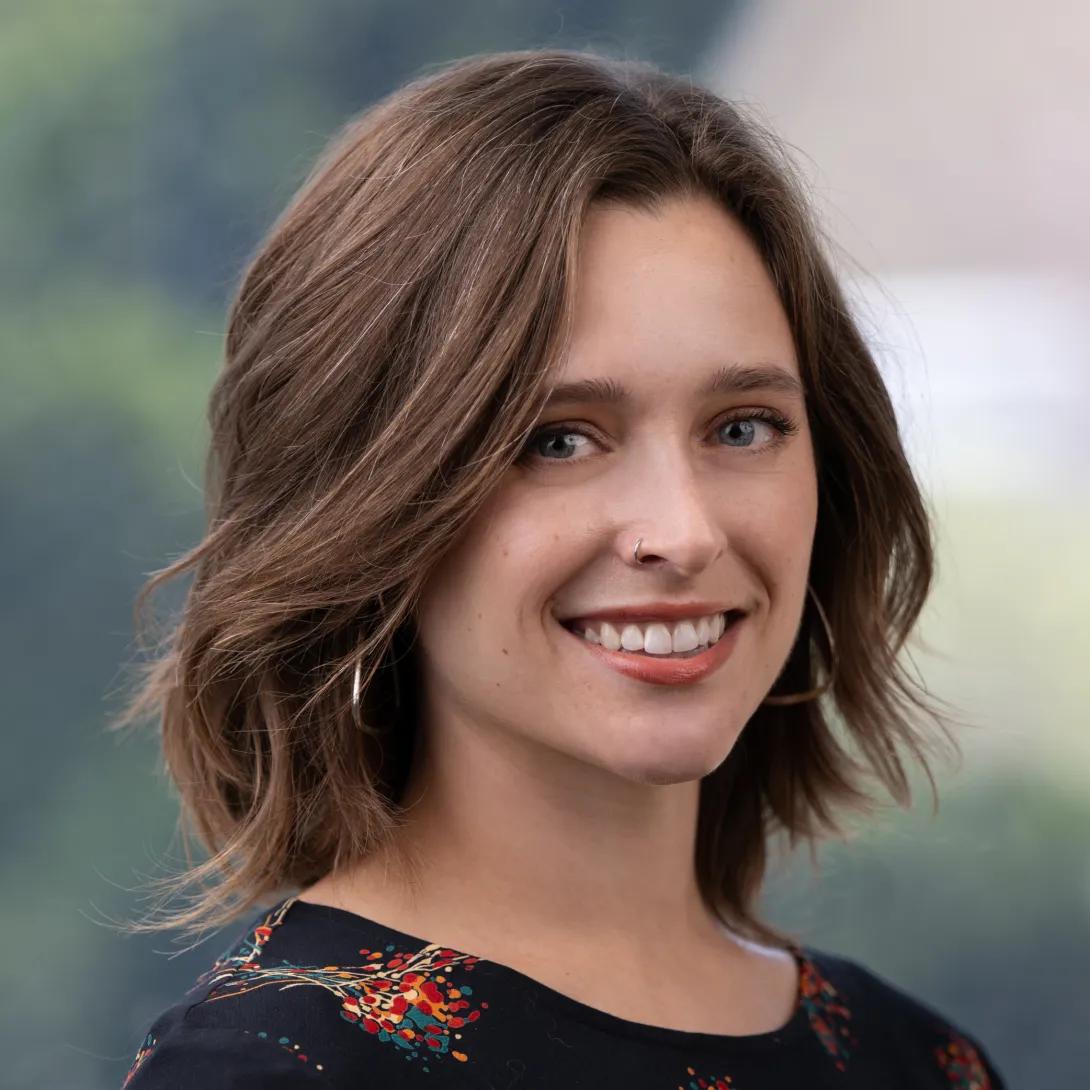 Head-and-shoulders portrait of a smiling woman with shoulder-length wavy brown hair, a nose ring, and hoop earrings, wearing a dark floral top against a softly blurred green background.