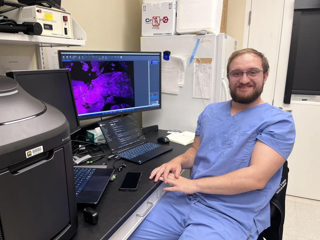 Man in blue scrubs smiling next to computer with image of stained cells.