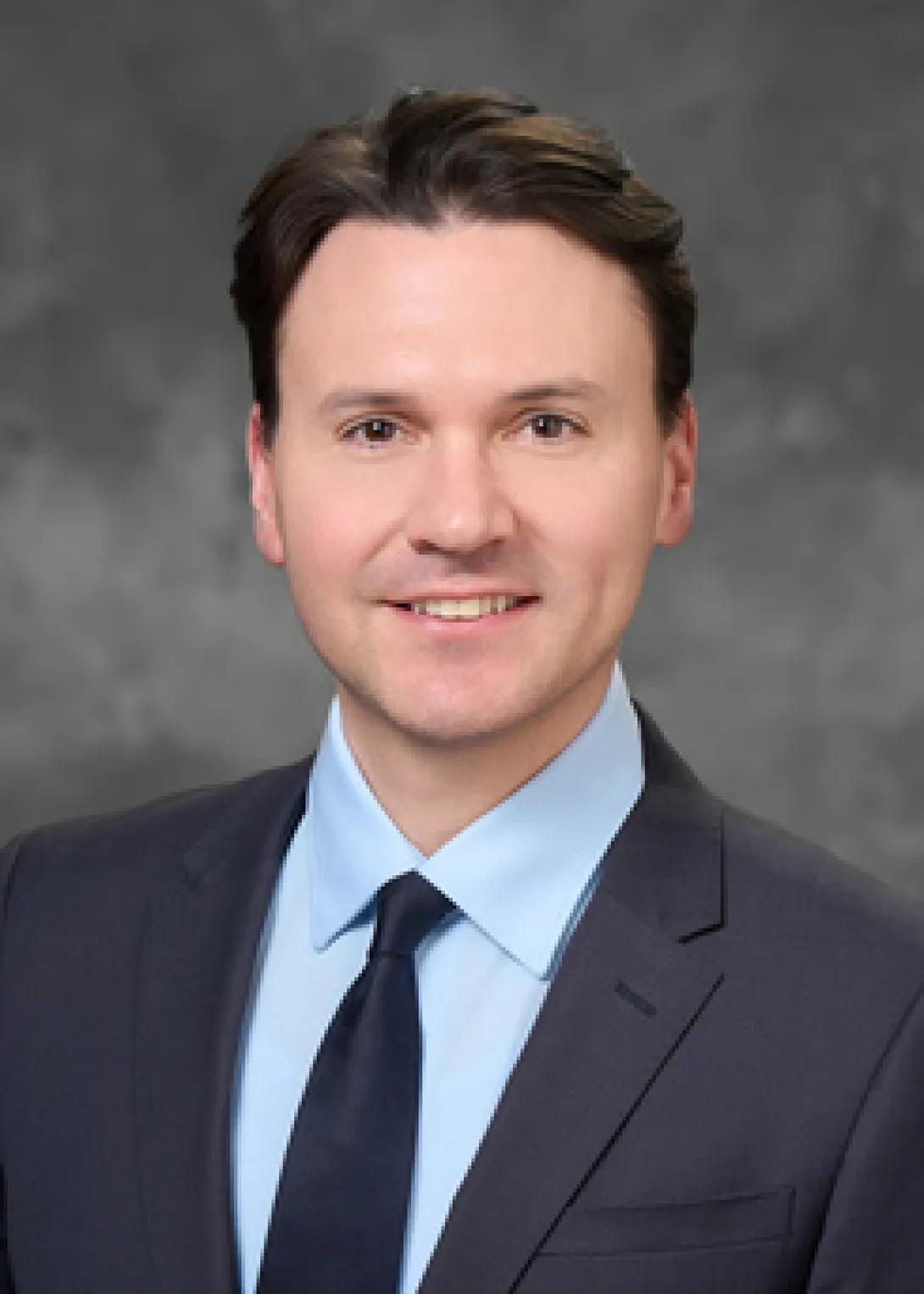 Headshot of person with short brown hair, smiling in a navy suit, light blue shirt and dark navy neck tie.