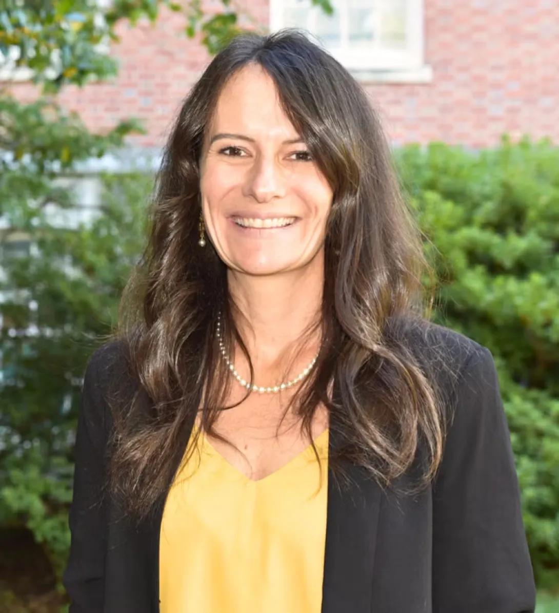 Headshot of person with long dark hair wearing a pearl necklace, yellow v-neck top and black blazer standing outside with trees behind them.