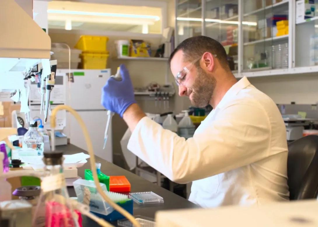 Man in white coat and safety goggles pipetting at a lab bench.