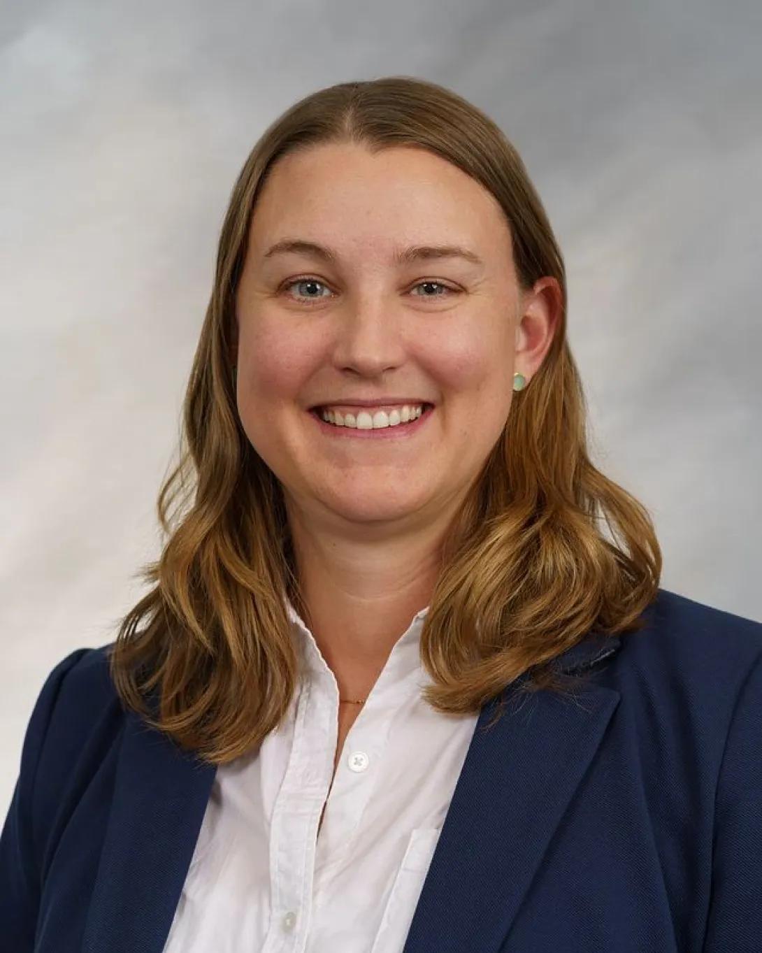 Headshot image of person smiling with longer brown hair, wearing a white top and navy blazer