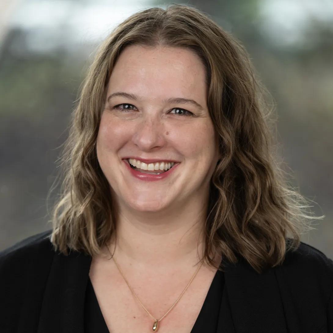 Professional headshot of a smiling woman with shoulder-length wavy brown hair, wearing a black blazer and gold pendant necklace against a softly blurred gray background.