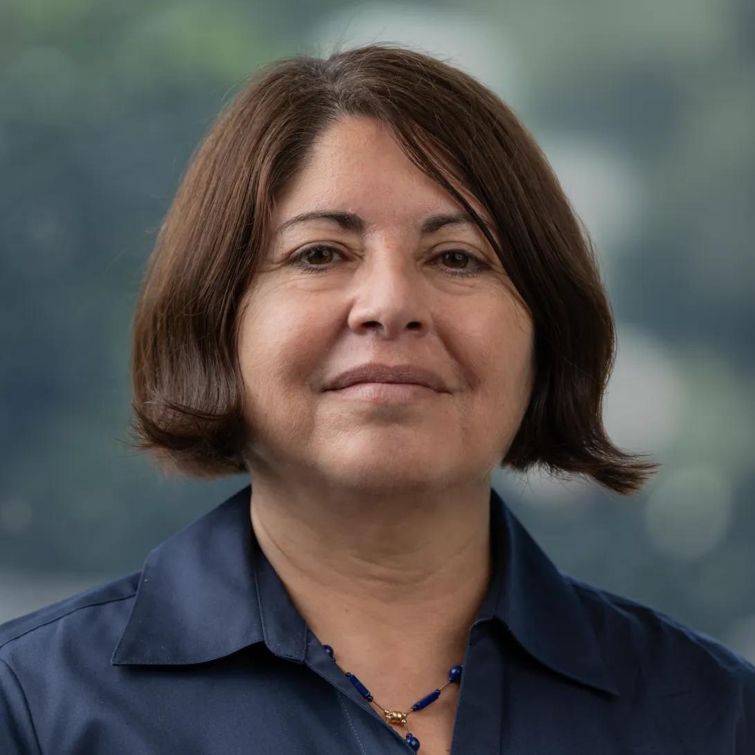 Professional headshot of a woman with short brown hair wearing a navy collared shirt and blue necklace against a softly blurred background.