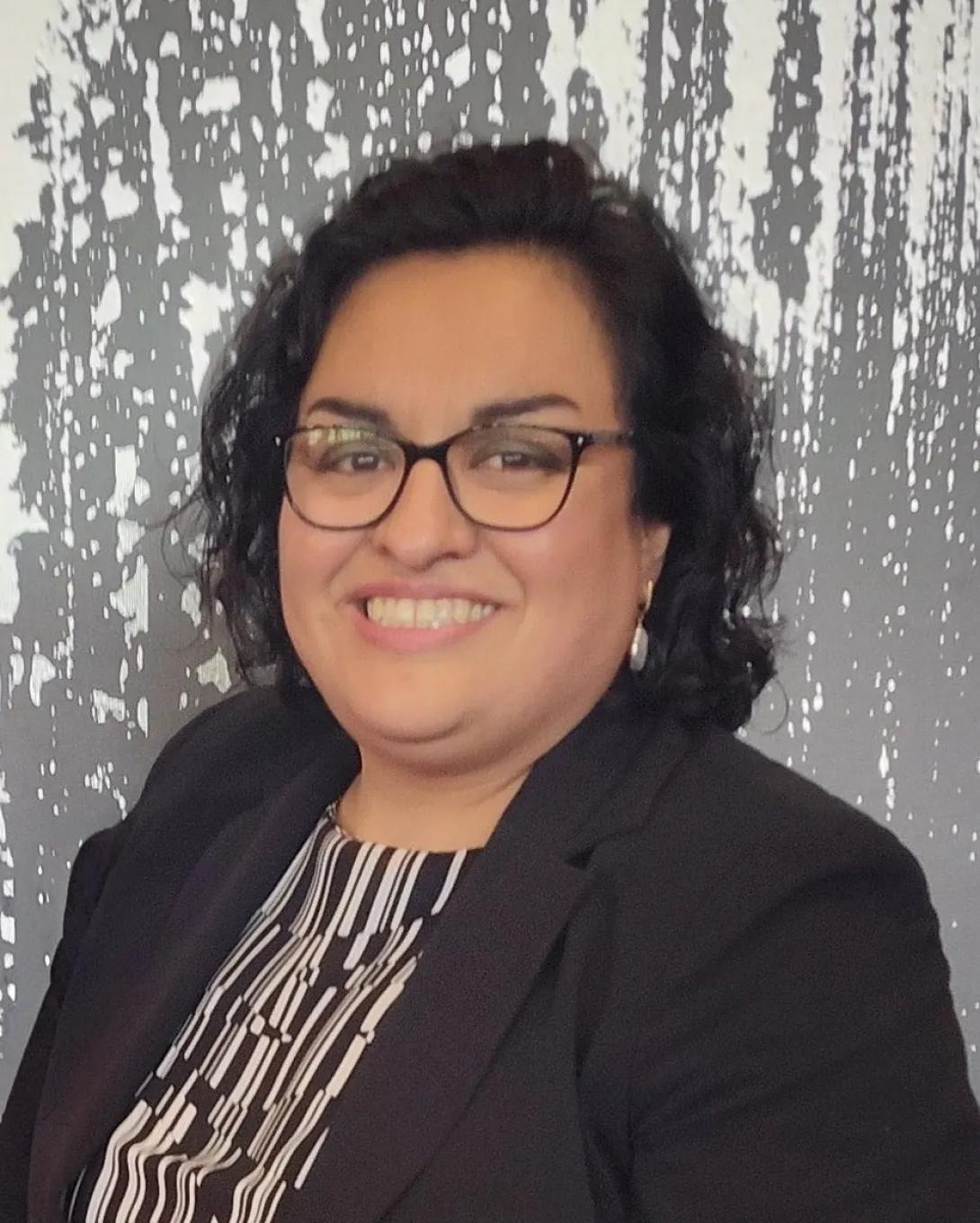 Maria Flores: a woman with short black hair and glasses smiles wearing a black blazer and black and white shirt in front of a gray and white background