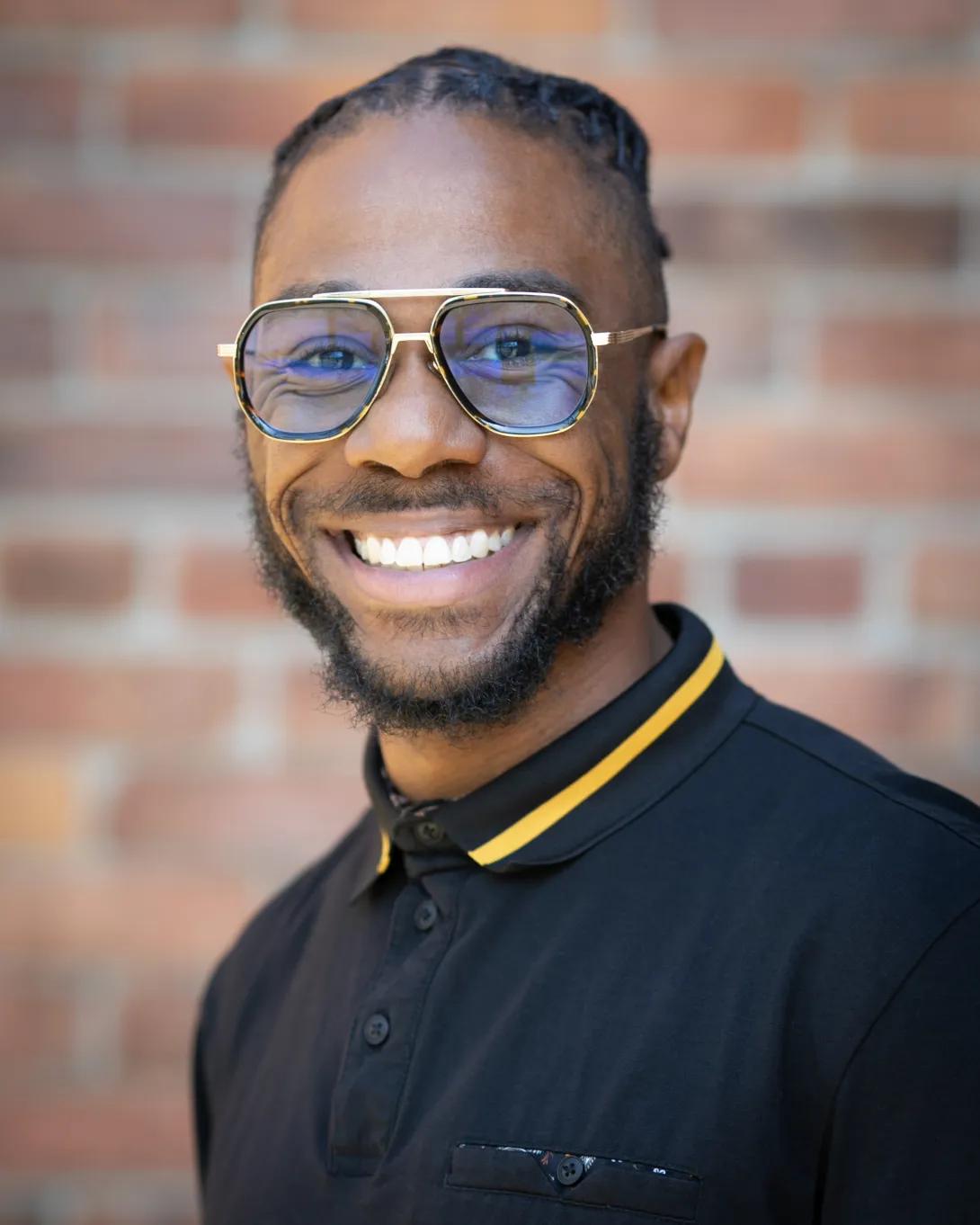 Teddy Dorsette III smiling at the camera, wearing glasses and a black polo shirt, with a brick wall in the background.