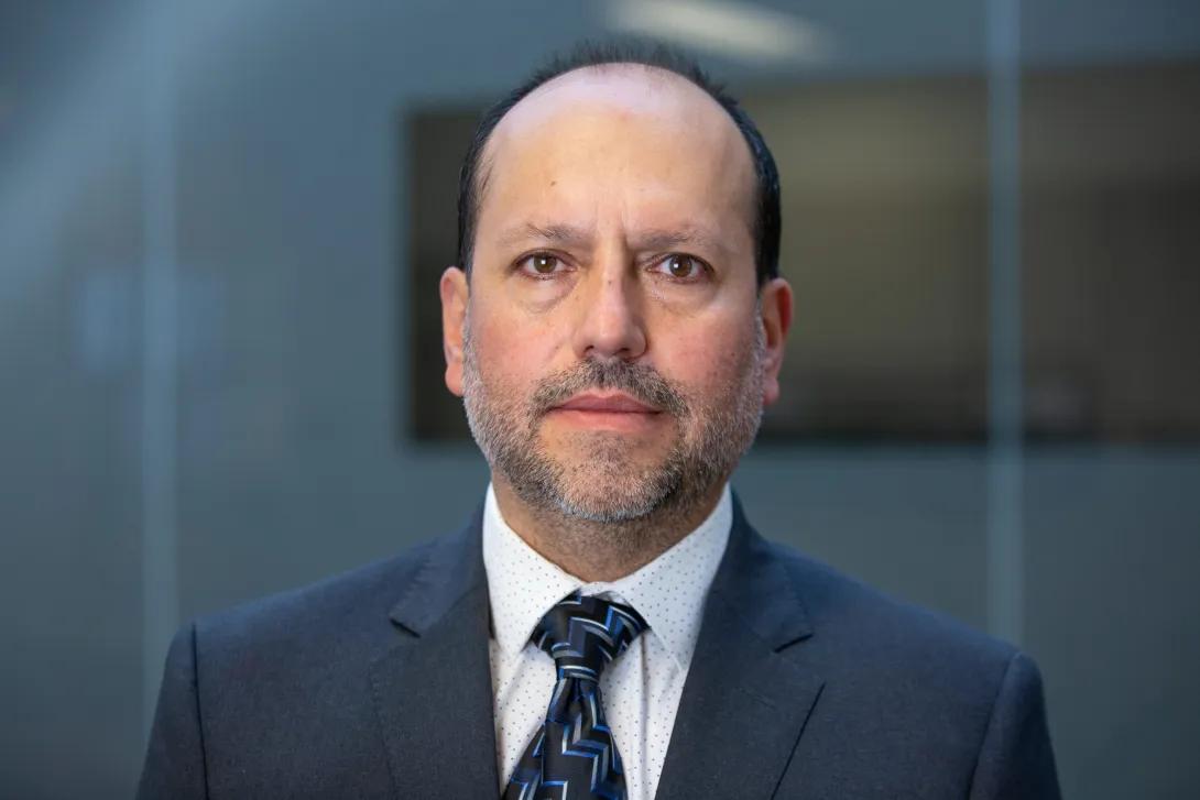 Man in front of a blurred background wearing a navy suit, white shirt, and navy tie